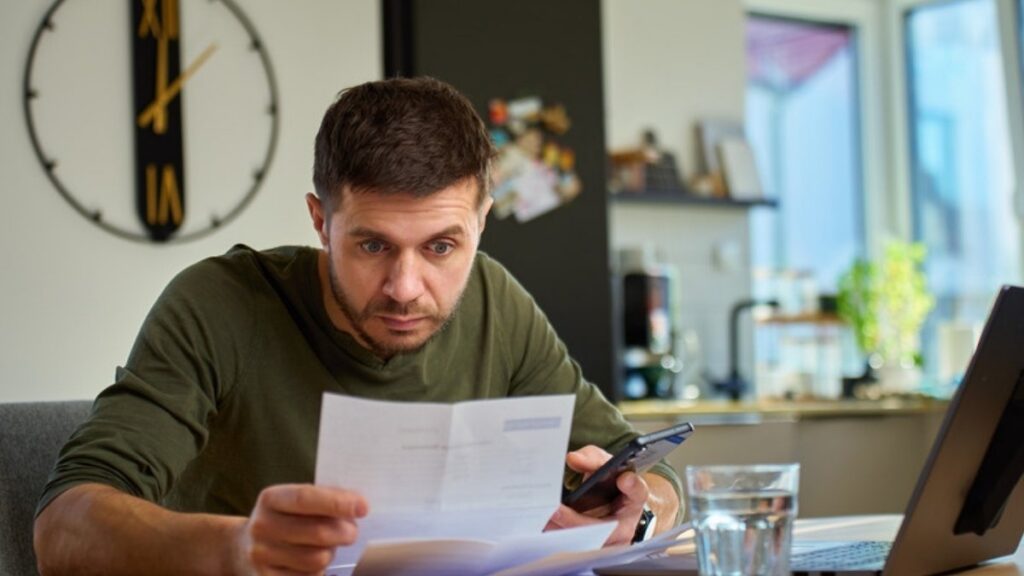 A man frowning over a letter at his kitchen table with his phone in hand