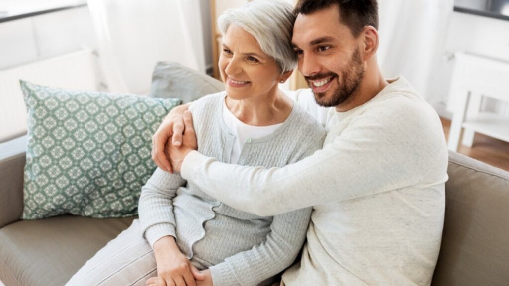 An older mother and her adult son sitting close together on the living room sofa