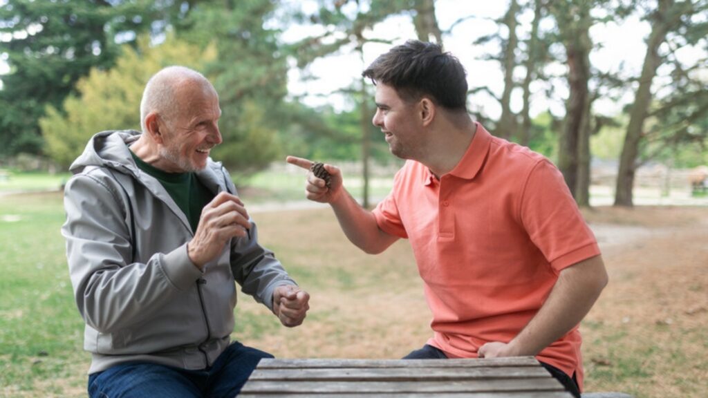 An older father and his adult son with Down syndrome laughing at a park picnic table, a family planning how a special needs trust protects SSI eligibility