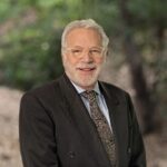 Conservatorship attorney Howard Gold in a dark suit, patterned tie and glasses, with green trees and a wooded path behind him