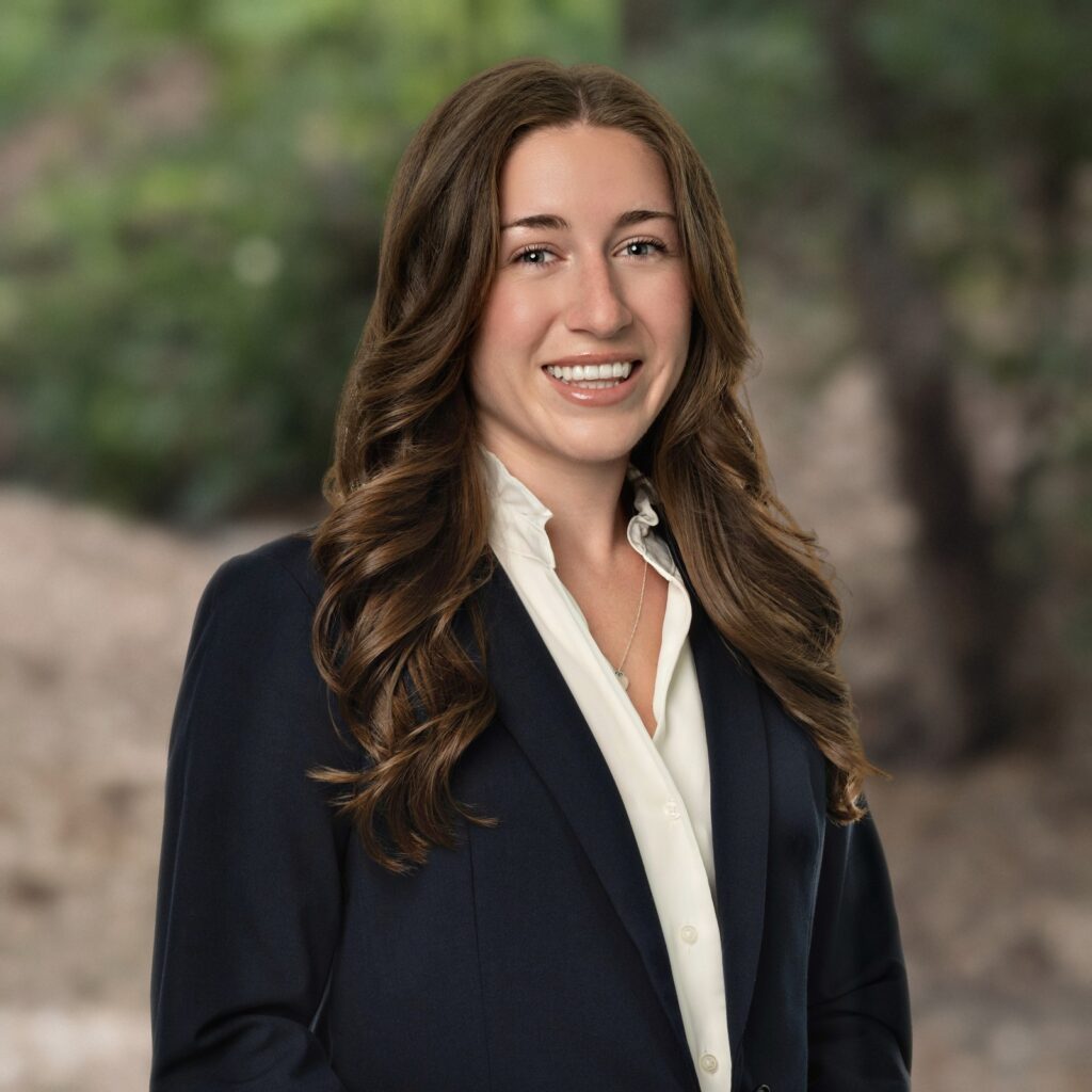 Estate planning attorney Megan Lee in a navy blazer and white blouse, smiling with soft green woods behind her