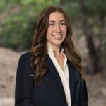 Estate planning attorney Megan Lee in a navy blazer and white blouse, smiling with soft green woods behind her